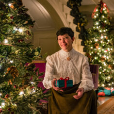 Person in a white shirt holding a wrapped gift next to a lit Christmas tree.